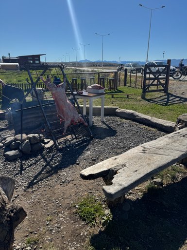 Ein Grillplatz im Freien mit einem großen Stück Fleisch, das über einem offenen Feuer hängt, umgeben von einer malerischen Landschaft.