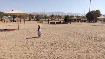 Ein kleines Mädchen mit langen Haaren läuft fröhlich über einen sandigen Platz, umgeben von Palmen und Spielgeräten.