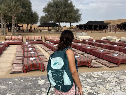 Eine Person mit einem Rucksack steht auf einem gepflasterten Weg und blickt auf eine Wüstenlandschaft mit Zelten.