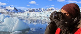 Ein Fotograf in warmer Kleidung hält eine Kamera, während er die beeindruckende arktische Landschaft mit Eisbergen und Bergen festhält.