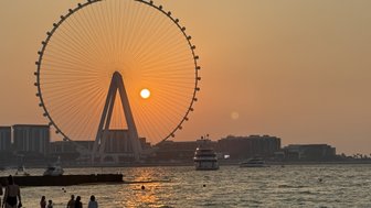 Ein Sonnenuntergang hinter einem großen Riesenrad, während Menschen am Strand spazieren und Boote im Wasser schwimmen.