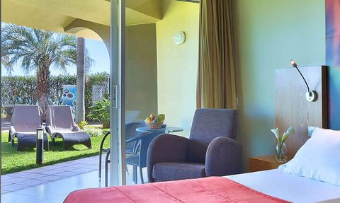 Ein helles Hotelzimmer mit einem roten Bettüberwurf, Blick auf einen Garten mit Liegen und einem Tisch im Freien.
