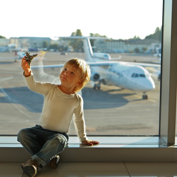 Ein kleiner Junge sitzt am Fenster eines Flughafens und hält ein Spielzeugflugzeug in der Hand, während er nach draußen schaut.