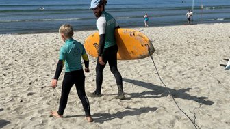 Ein Surflehrer und ein Junge gehen am Strand entlang, während der Lehrer ein großes, orangefarbenes Surfbrett trägt.