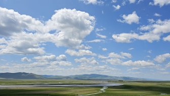 Eine weite, grüne Landschaft unter einem strahlend blauen Himmel mit vielen weißen Wolken und sanften Hügeln im Hintergrund.