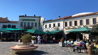 Ein belebter Platz mit Cafés und Restaurants, umgeben von historischen Gebäuden und Sonnenschirmen, unter strahlend blauem Himmel.