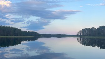 Ein ruhiger See spiegelt den Himmel mit sanften Wolken und einem Hauch von Abendlicht wider, umgeben von dichten Wäldern.