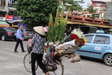 Eine Frau mit einem traditionellen conical Hut fährt mit ihrem Fahrrad, beladen mit bunten Blumen, durch eine belebte Straße.