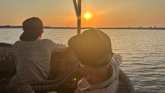 Zwei Personen sitzen auf einer Terrasse und beobachten den Sonnenuntergang über dem Wasser, umgeben von einer friedlichen Landschaft.