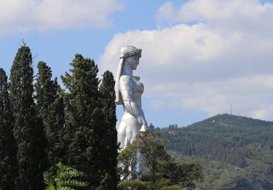 Die Statue einer Frau mit einer Krone steht majestätisch zwischen hohen Bäumen, umgeben von sanften Hügeln und blauem Himmel.