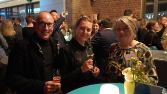 Drei Personen lächeln fröhlich, während sie Gläser mit Getränken in einer festlichen Umgebung halten.