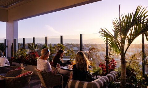 Drei Personen sitzen entspannt in einer Dachterrassenbar, umgeben von Pflanzen und genießen den Sonnenuntergang über der Stadt.