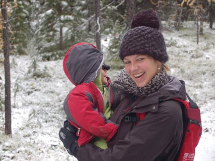 Eine lächelnde Frau hält ein kleines Kind in einem roten Anorak, umgeben von schneebedeckten Bäumen.
