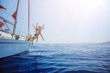 Drei fröhliche Personen springen von einem Segelboot ins glitzernde blaue Wasser, umgeben von strahlendem Sonnenschein.