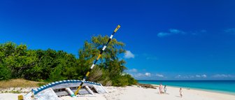 Ein verlassenes Boot liegt auf dem weißen Sandstrand, während eine Familie am Ufer entlang spaziert und die Sonne genießt.