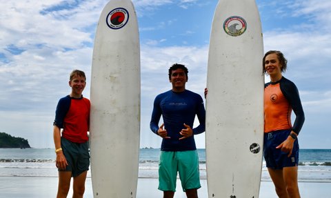 Drei junge Surfer stehen mit ihren Brettern am Strand von Playa Samara, umgeben von sanften Wellen und blauem Himmel.