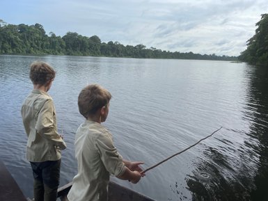 Zwei Jungen stehen am Ufer eines ruhigen Gewässers und versuchen, mit einem Stock zu fischen, umgeben von üppigem Grün.