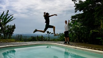 Ein Junge springt fröhlich in einen glasklaren Pool, während ein Freund ihm begeistert zuschaut und den Daumen hebt.