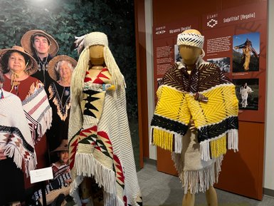 Zwei kunstvoll gekleidete Figuren in traditioneller Regalia stehen vor einer Wand mit kulturellen Informationen.