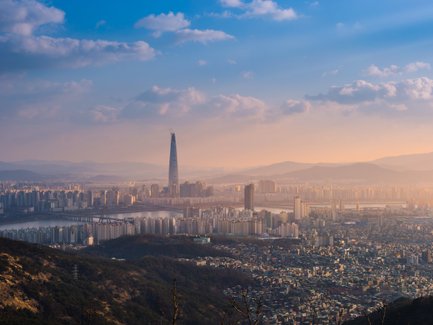 Die Skyline von Seoul bei Sonnenuntergang, mit dem markanten Lotte World Tower, umgeben von sanften Hügeln und Wolken.