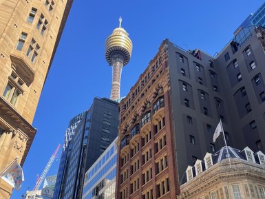 Das Bild zeigt eine belebte Stadtansicht, dominiert von modernen und historischen Gebäuden unter klarem, blauem Himmel.