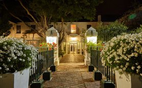 Der Eingangsbereich des Windsor Hotels in Hermanus ist nachts beleuchtet, umgeben von blühenden Blumen und grünen Bäumen.