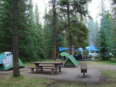 Ein Campingplatz im Wald mit grünen Zelten, einem Picknicktisch und blauen Planen, umgeben von hohen Bäumen.