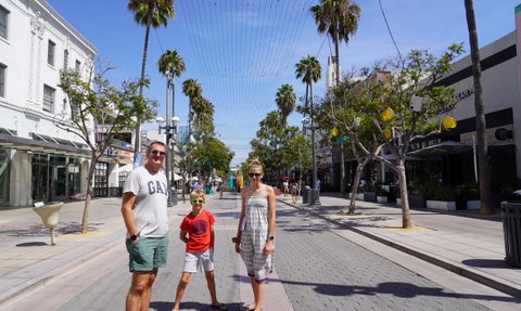 Eine Familie steht fröhlich auf der Santa Monica Promenade, umgeben von Palmen und Geschäften unter blauem Himmel.