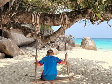 Ein Junge sitzt auf einer Holzschaukel, umgeben von einem malerischen Strand und sanften Wellen im Hintergrund.