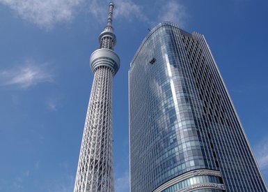 Der Tokyo Skytree erhebt sich majestätisch über die moderne Architektur eines angrenzenden Wolkenkratzers unter blauem Himmel.