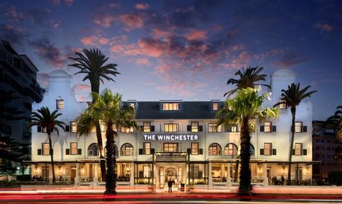 Das Winchester Hotel strahlt in der Dämmerung, umgeben von Palmen und einem farbenfrohen Himmel.