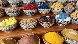 Bunte Schalen mit verschiedenen Gewürzen und getrockneten Blumen auf einem Holztisch, umgeben von einer grünen Pflanzenwand.