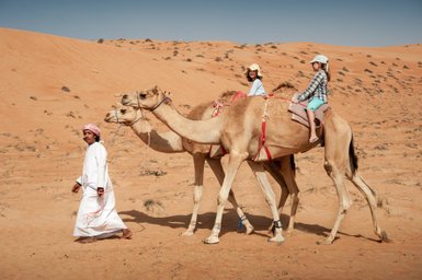 Kinder reiten gemeinsam mit einem Einheimischen auf Kamelen – Oman Reise mit Kindern