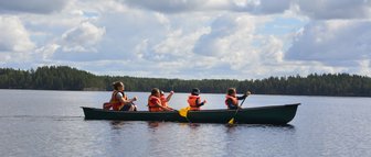 Eine Gruppe von vier Personen paddelt in einem Kanu auf einem ruhigen See, umgeben von üppigem Wald und bewölktem Himmel.