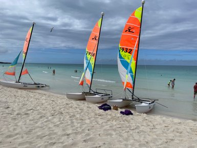 Drei bunte Katamarane stehen am Strand, während Menschen im klaren Wasser planschen und die Wolken am Himmel ziehen.