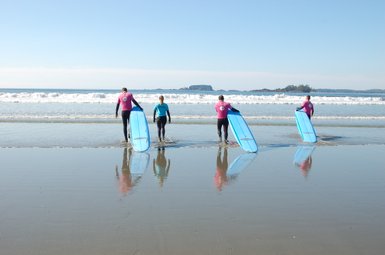 Vier Surfer gehen mit ihren blauen Surfbrettern ins Wasser, während die Wellen sanft am Strand brechen.