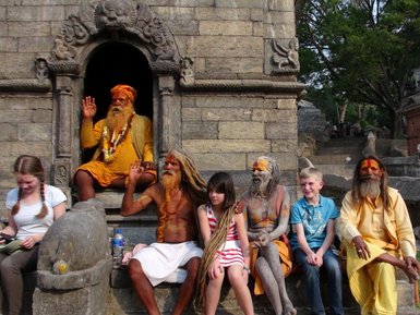 Eine Gruppe von Sadhu-Mönchen sitzt entspannt auf einer Steinbank, während Kinder und Erwachsene in der Nähe lächeln.