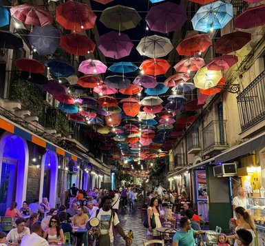 Bunte Regenschirme hängen über einer belebten Gasse, in der Menschen an Tischen sitzen und sich unterhalten.