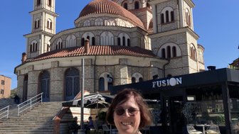 Eine Frau mit Brille lächelt vor einer beeindruckenden Kirche mit roten Kuppeln und Türmen unter klarem blauen Himmel.