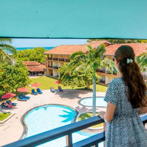 Eine Frau mit langen Haaren steht auf einem Balkon und blickt auf einen Pool und die üppige Gartenlandschaft.