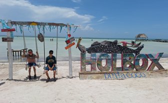 Zwei Kinder schwingen fröhlich auf einer Schaukel am Strand von Holbox, umgeben von klarem Wasser und einem blauen Himmel.