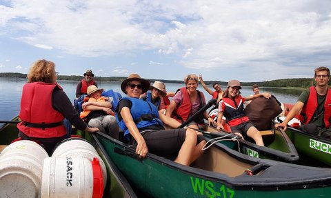 Eine Gruppe von Menschen in Kanus genießt einen sonnigen Tag auf einem ruhigen See, umgeben von grüner Natur.