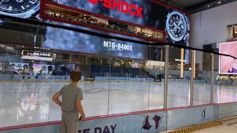 Ein Junge steht am Rand einer Eislaufbahn und beobachtet die leere Eisfläche, während große Bildschirme Werbung zeigen.