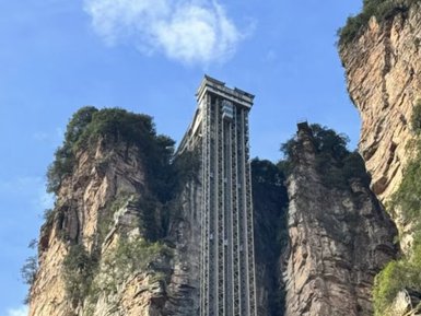 Ein beeindruckender gläserner Aufzug ragt zwischen steilen Felsen in den Himmel, umgeben von üppigem Grün und klarem blauen Himmel.