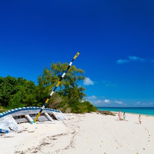Ein verlassenes Boot liegt auf dem weißen Sandstrand, während eine Familie am Ufer entlang spaziert und die Sonne genießt.