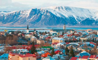 Blick auf Reykjavik mit bunten Häusern, umgeben von schneebedeckten Bergen und einem ruhigen Wasser im Hintergrund.