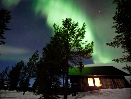 Ein Holzhaus steht in einer verschneiten Landschaft, während die Nordlichter in leuchtendem Grün darüber tanzen.
