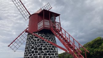 Eine rote Windmühle mit Steinstütze steht vor einem bewölkten Himmel, während eine Person auf der Treppe steht.