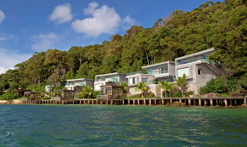 Moderne Strandvillen am Wasser, umgeben von üppigem Grün und klarem Himmel, ideal für einen entspannten Urlaub.