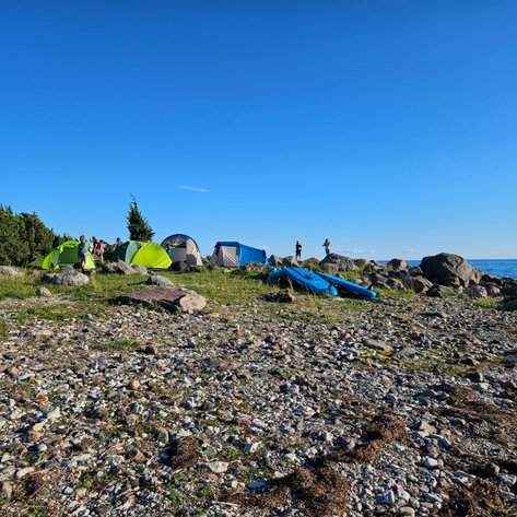 Eine Gruppe von Zelten steht am Ufer, umgeben von Steinen und Gras, während das Wasser ruhig im Hintergrund glitzert.